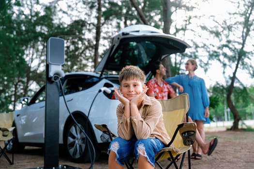 Little boy portrait sitting on camping chair with his family in background. Road trip travel with alternative energy charging station for eco-friendly car concept. Perpetual