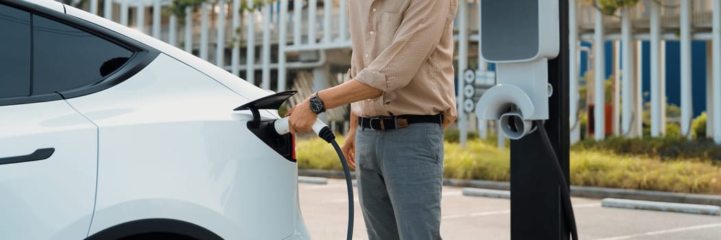 Young man recharge electric car's battery from charging station in city commercial parking lot. Rechargeable EV car for sustainable environmental friendly urban travel. Panorama Expedient
