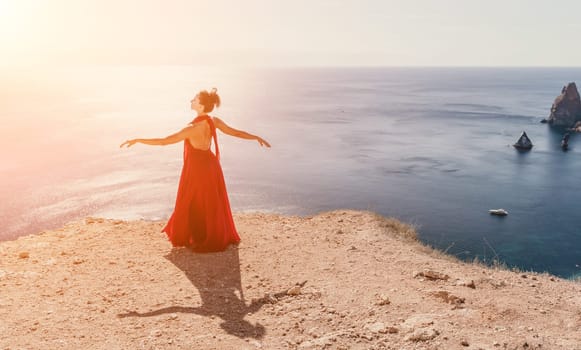 Side view a Young beautiful sensual woman in a red long dress posing on a rock high above the sea during sunrise. Girl on the nature on blue sky background. Fashion photo.