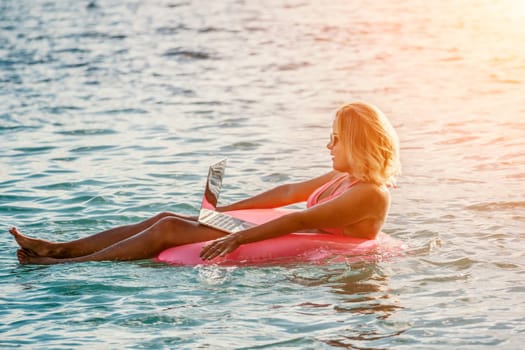 Woman works on laptop in sea. Freelancer, young blond woman in sunglases floating on an inflatable big pink donut with a laptop in the sea at sunset. Freelance, travel and holidays concept