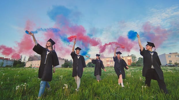 Graduates in costume walk with a smoky multi-colored smoke at sunset.