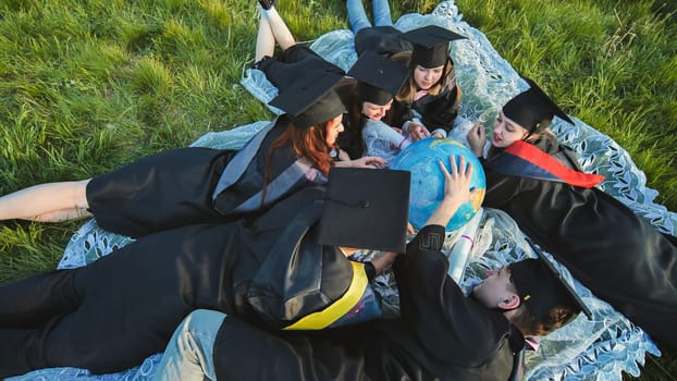 Graduate students in black robes study a globe on the grass