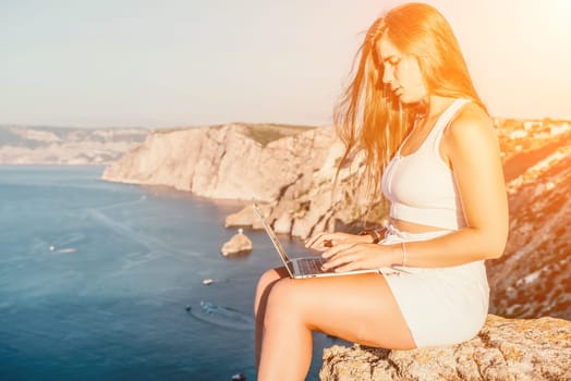 Successful business woman in yellow hat working on laptop by the sea. Pretty lady typing on computer at summer day outdoors. Freelance, travel and holidays concept.