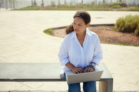 Confident pensive business woman thinking on new startup project, online working on laptop, sitting on bench outdoors and dreamily looking to the side. Remote job. People. Career. Distance work