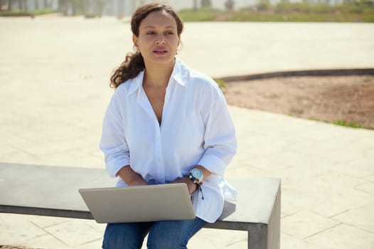 Confident young business woman sitting outdoors with laptop on knees. Online messaging. Remote work. People. Job. Online communication. Connection