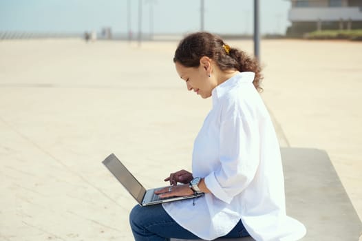 Side portrait of a confident young business woman sitting outdoors with laptop on knees. Online messaging. Remote work. People. Job. Online communication. Connection. Telecommuting. Freelancer