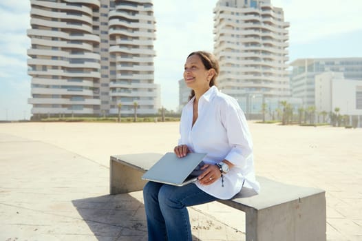 Authentic portrait of a happy young adult multi ethnic businesswoman, holding laptop, sitting on a bench against cityscape background. People. Career. Recruitment. Online business. Remote job