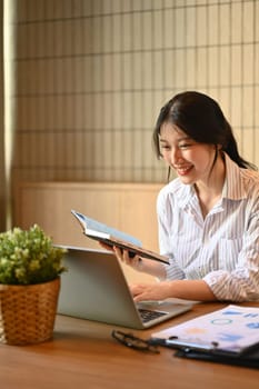 Beautiful young businesswoman holding notepad and checking information on laptop computer.