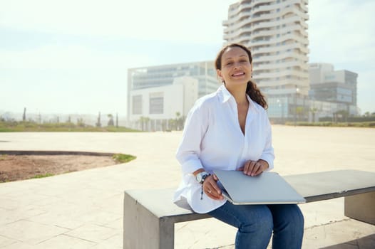 Authentic portrait of a young adult multi ethnic woman, business lady, freelancer, entrepreneur, developer sitting on a bench against cityscape background and looking at camera, enjoying remote job