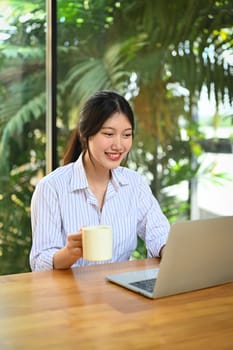 Happy Asian female office worker drinking coffee and working on laptop computer.