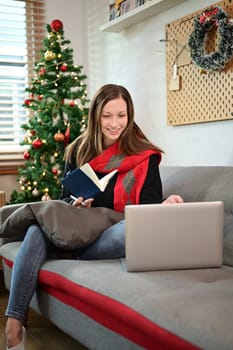 Smiling young woman having video call via laptop while sitting on couch in living room decorated for Christmas.