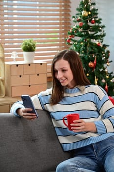 Happy caucasian woman using smartphone while sitting on couch against Christmas tree with lights.