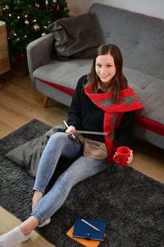 Above view of young woman sitting on floor against Christmas tree and browsing internet on digital tablet.