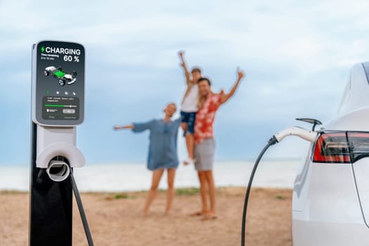 Alternative family vacation trip traveling by the beach with electric car recharging battery from EV charging station with blurred cheerful and happy family enjoying the seascape background. Perpetual