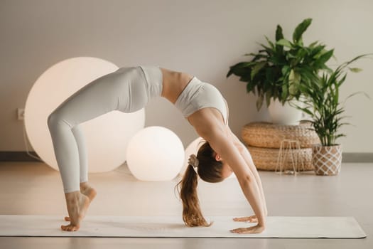 A girl in white clothes does yoga standing on the bridge on a mat indoors.