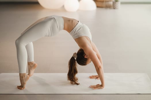 A girl in white clothes does yoga standing on the bridge on a mat indoors.