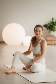 Portrait of a girl in white clothes sitting on a mat before doing Yoga indoors.