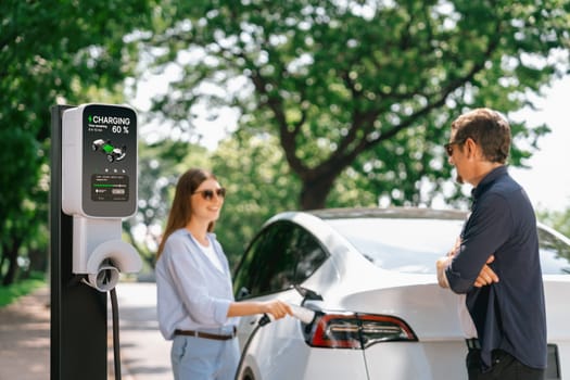 Lovely young couple wearing sun glasses recharging battery for electric car during road trip travel EV car in natural forest or national park. Eco friendly travel during vacation and holiday. Exalt
