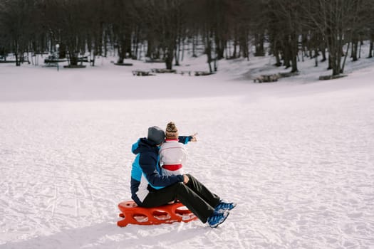 Dad with a small child sits on a sled on a snowy plain and points towards the forest. Back view. High quality photo