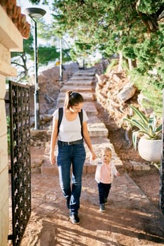 Mom and little girl walk along a paved path with steps in the park, holding hands. High quality photo