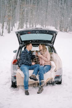 Smiling man and woman with cups in their hands are sitting in the trunk of a car in a snowy forest. High quality photo