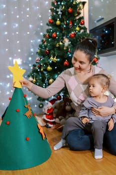 Little girl sits on her mother lap and watches as she sets a star on a felt Christmas tree. High quality photo