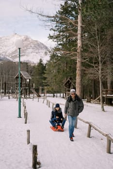 Smiling dad carries mom and little boy on a sleigh along a path in the snowy forest. High quality photo