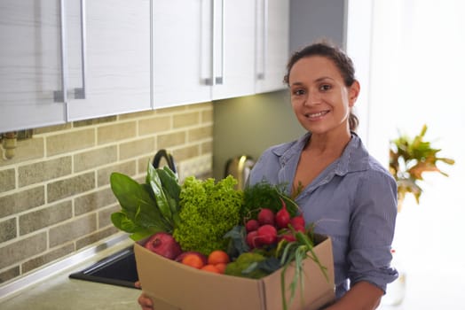 Smiling multi ethnic brunette, pleasant housewife holding a cardboard with fresh veggies, fruits and greens, smiling looking at camera, standing by kitchen counter at home. People. Healthy food