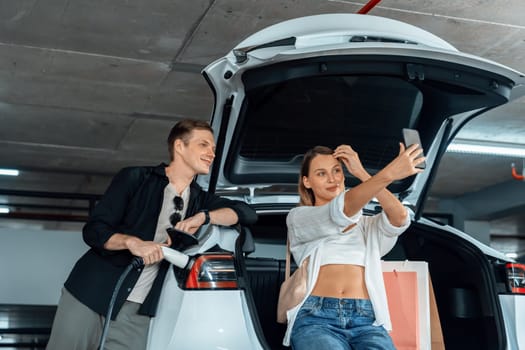 Young couple travel with EV electric car to shopping center parking lot charging in downtown city showing urban sustainability lifestyle by green clean rechargeable energy of electric vehicle innards