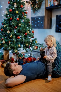 Little smiling girl with a reflex camera in her hands sits on her dad stomach and looks at him near the Christmas tree. High quality photo