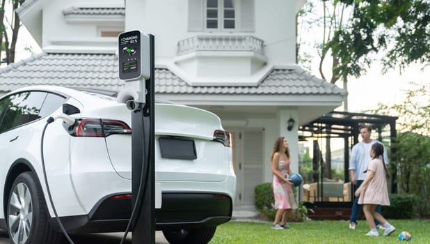 Focus electric vehicle recharge from home charging station on blur background of happy and playful family playing together. EV car using alternative and sustainable energy for better future.Synchronos