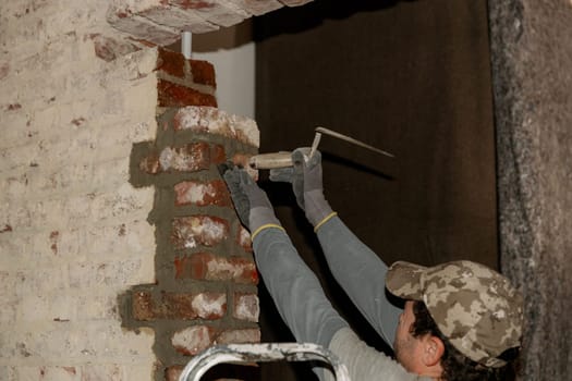 One young Caucasian man lays bricks on fresh cement in a doorway, tapping with the handle of a spatula, close-up side view.