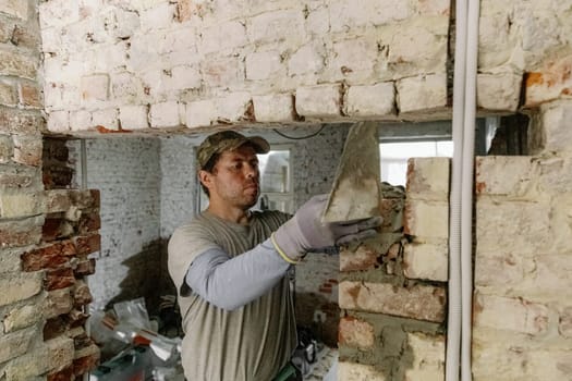 One young Caucasian man lays bricks on fresh cement in a doorway, leveling it with his hands and standing on a stepladder to the right in an old house, close-up side view.