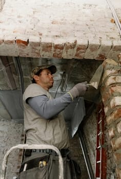 One young Caucasian man spreads fresh cement onto bricks using a spatula, leveling it and standing during the day on the right on a stepladder in an old house, close-up view from below.