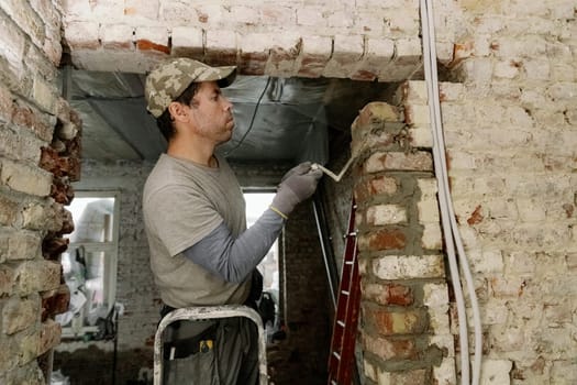 One young Caucasian man inspects a brick laid on fresh cement, holding a spatula in his hands and standing on a stepladder to the right during the day in an old house, close-up view from below.