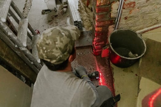 One young Caucasian man in casual clothes squats and measures fresh brickwork in a doorway in an old house using a laser level, close-up side view.