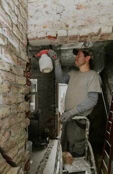 One young handsome Caucasian happy brunette man in casual clothes stands on a stepladder and sprays water from a spray bottle onto bricks in a doorway under the ceiling in an old house, close-up view from below.