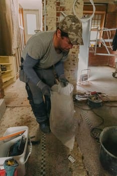 One young brunette Caucasian man in a gray casual work uniform holds a white bag of construction waste while standing in an old house, a destroyed house, where renovations are underway, close-up side view.