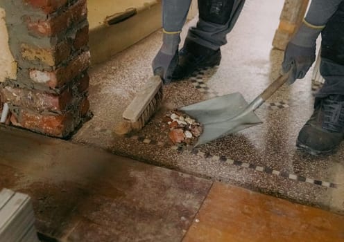 One young Caucasian man, unrecognizable in a gray casual work uniform, sweeps construction debris into a dustpan with a broom after laying bricks in a doorway, standing in an old ruined house, close-up side view.