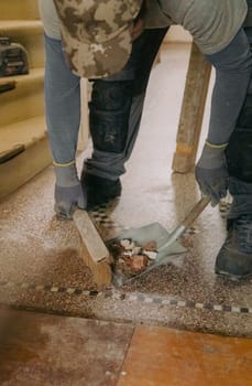 One young Caucasian man, unrecognizable in a gray casual work uniform, sweeps construction debris into a dustpan with a broom after laying bricks in a doorway, standing at an angle in an old ruined house, close-up side view.