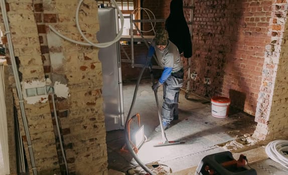 One young Caucasian man in a gray casual work uniform vacuums up construction debris after laying bricks in a room where renovations are underway, standing under the light of a lantern in an old ruined house, close-up side view.