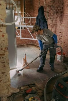 One young Caucasian man in a gray casual work uniform vacuums up construction debris after laying bricks in a room where renovations are underway, standing sideways under the light of a lantern in an old ruined house, close-up side view.