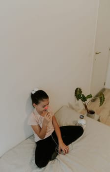 One beautiful Caucasian brunette girl with a happy smile, collected hair and in a pink T-shirt treats the right nasal passage with a device with infrared light, sitting on the bed, cross-legged, leaning on a white wall, close-up view from above.