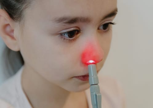 One beautiful Caucasian brunette girl with her hair tied up and wearing a pink T-shirt is treating her right nasal passage with an infrared light machine, sitting against a white wall, side view, very close-up.