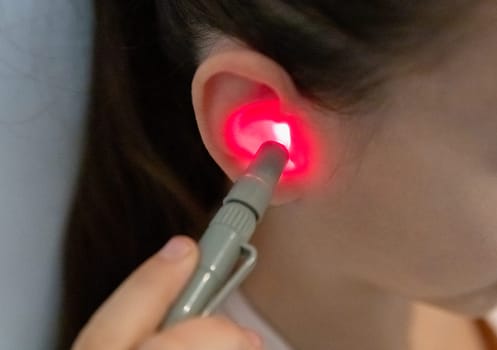 One beautiful Caucasian brunette girl with a happy smile, collected hair and in a pink T-shirt treats her right ear with an infrared light device, sitting on a bed at home near a white wall, top-side view very close-up.