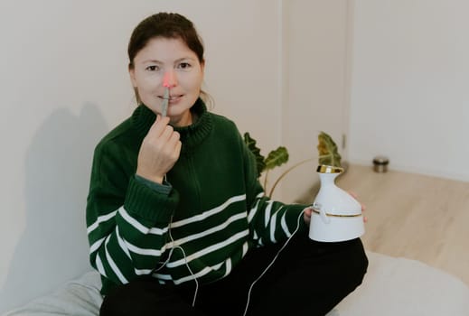 One beautiful Caucasian brunette girl with collected hair in a green sweater treats the right nasal passage with an apparatus with infrared light, sitting on a bed against a white wall, close-up side view from above.