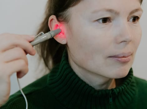 One beautiful Caucasian brunette girl with collected hair in a green sweater treats her right ear with an infrared light device, sitting on a bed against a white wall, side view very close-up.