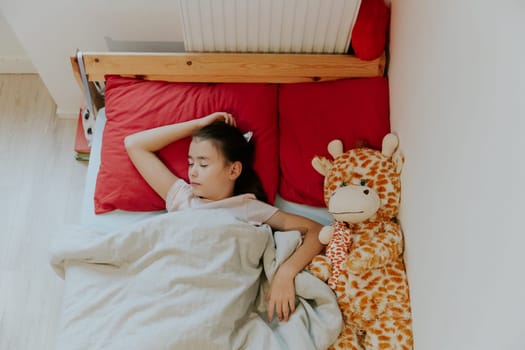 One beautiful little Caucasian brunette girl sleeps sweetly in a wooden bed on a red pillow with a soft toy giraffe, covered with a gray blanket, flat lay close-up.