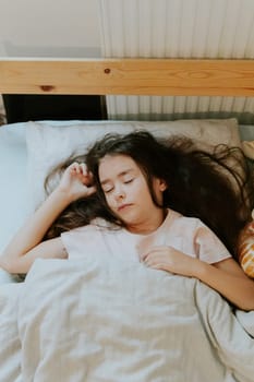 One beautiful little Caucasian brunette girl sleeps sweetly in a wooden bed with her long hair flowing on a pillow covered with a gray blanket, close-up top view.