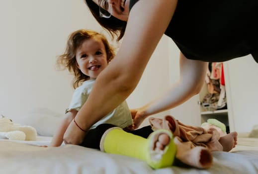 One Caucasian mother with a smile on her face is changing her cheerful baby daughter with a cast on her leg and showing her tongue in the morning in the room on the bed, side view close-up.